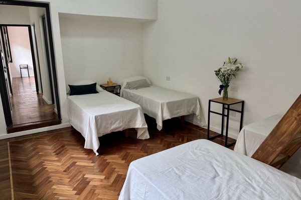 Amplio dormitorio con varias camas, altillo accesible por escalera de madera y suelo de parquet. Paredes blancas y ventanas con luz natural. Mobiliario funcional incluye mesitas de noche y perchero.