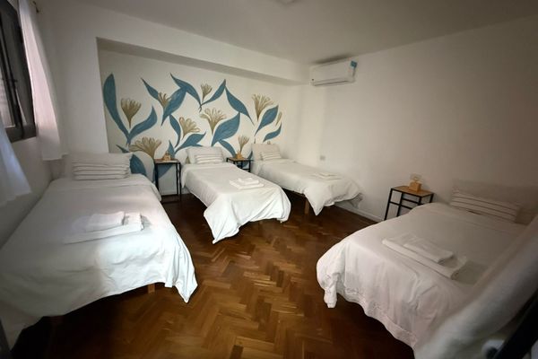 Dormitorio luminoso con varias camas, veladores con lámparas cálidas y detalles naturales. Vista desde una pequeña abertura decorada con plantas que aporta un toque acogedor y relajado. Ideal para descansar en un ambiente sereno. Baño privado en Suite.