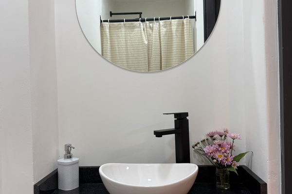 Baño compartido con ducha, equipado con sanitarios modernos y detalles minimalistas. Cuenta con lavamanos tipo bowl sobre estante negro, espejo redondo. Cortina de baño a rayas, tacho de residuos y toalla incluidos para mayor comodidad.