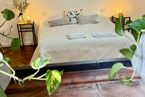 Dormitorio luminoso con cama queen, veladores con lámparas cálidas y detalles naturales. Vista desde una pequeña abertura decorada con plantas que aporta un toque acogedor y relajado. Ideal para descansar en un ambiente sereno.