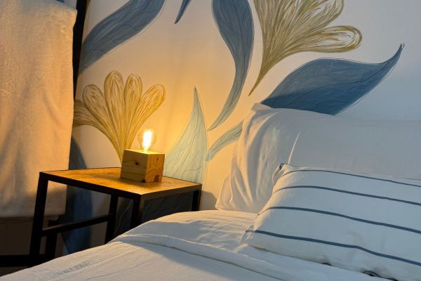 Mesa de luz cálida, mural de flores, cama blanca.Dormitorio luminoso con varias camas, veladores con lámparas cálidas y detalles naturales. Vista desde una pequeña abertura decorada con plantas que aporta un toque acogedor y relajado. Ideal para descansar