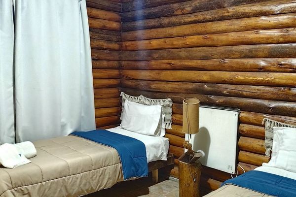Habitación rústica con dos camas individuales en cabaña de montaña ubicada en Potrerillos, Mendoza, sobre el Río Mendoza y la Ruta Internacional N°7 km 1108. Las camas están vestidas con acolchados beige, caminos de cama azul y almohadas blancas con fleco