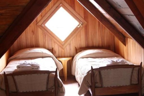 Interior de la habitación ‘Coihue’ en la hostería Wesley, con dos camas twin bajo un techo inclinado de madera y una ventana de rombo que deja entrar luz natural