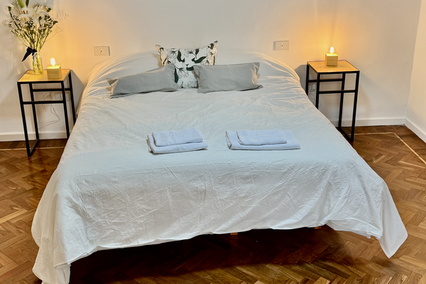 Dormitorio luminoso con cama queen, veladores con lámparas cálidas y detalles naturales. Vista desde una pequeña abertura decorada con plantas que aporta un toque acogedor y relajado. Ideal para descansar en un ambiente sereno.