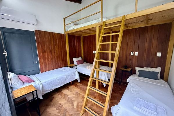 Habitación amplia con tres camas individuales en planta baja y una cama en entrepiso. La combinación de madera, techos altos y luz natural crea un ambiente cálido y confortable.