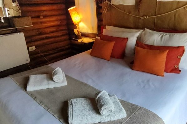 Dormitorio rústico y acogedor en cabaña de montaña ubicada en Potrerillos, Mendoza, sobre el Río Mendoza y la Ruta Internacional N°7 km 1108. La habitación presenta una decoración cálida con paredes de madera, cama matrimonial con sábanas blancas, manta b