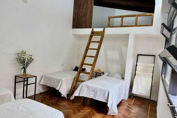 Amplio dormitorio con varias camas, altillo accesible por escalera de madera y suelo de parquet. Paredes blancas y ventanas con luz natural. Mobiliario funcional incluye mesitas de noche y perchero.